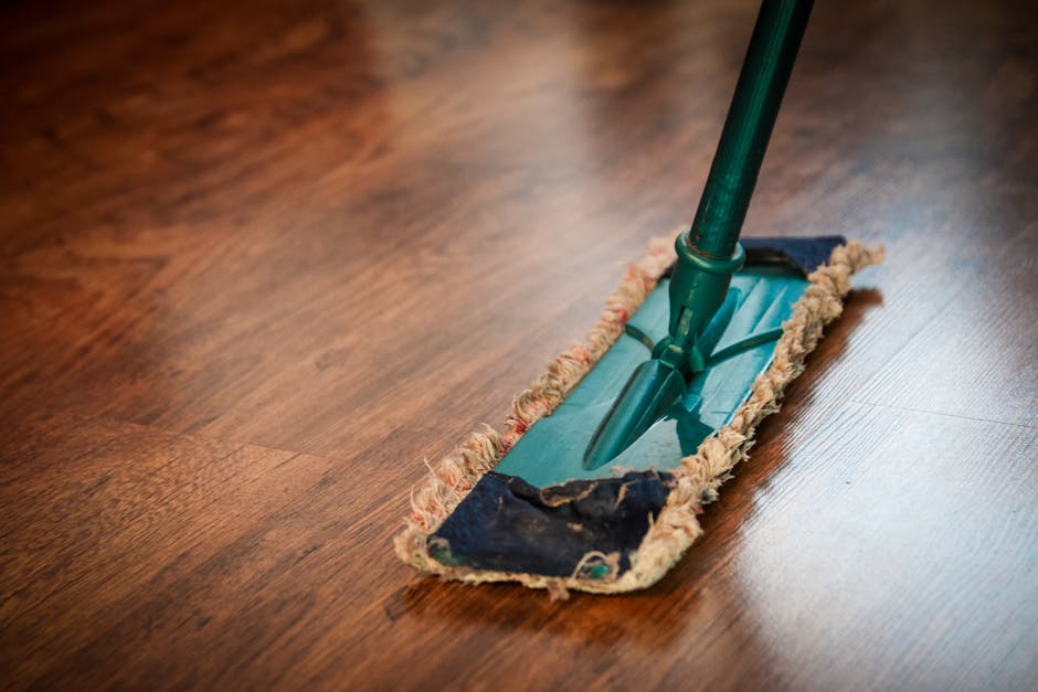 A close-up view of a wooden floor being cleaned with a green mop, featuring a flat mop head with a cloth cover that appears slightly worn. The surface of the floor is smooth, with a polished finish sh