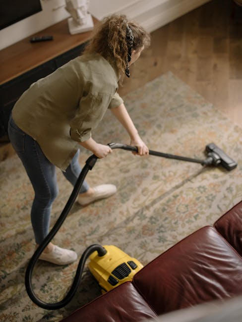 A woman with curly, light brown hair, wearing a beige sweater, jeans, and white shoes, is engaged in surface cleaning within a living room. She is vacuuming a large, patterned area rug with a yellow c
