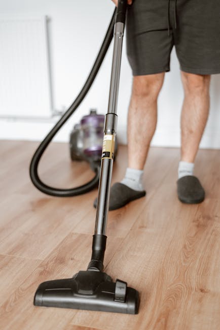 A woman with curly, light brown hair, wearing a beige sweater, jeans, and white shoes, is engaged in surface cleaning within a living room. She is vacuuming a large, patterned area rug with a yellow c